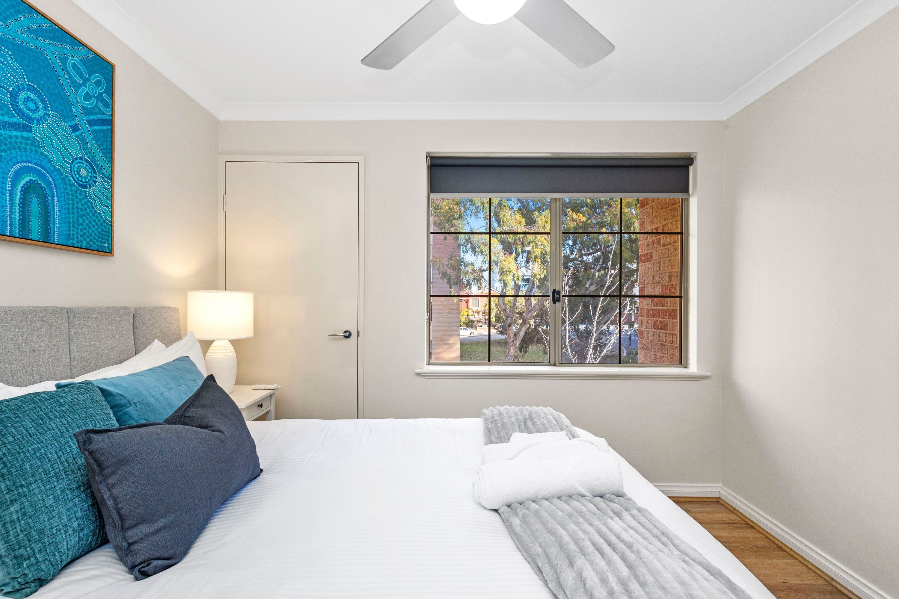 A bedroom with a bed, a window with a view, a ceiling fan, and a painting on the wall.