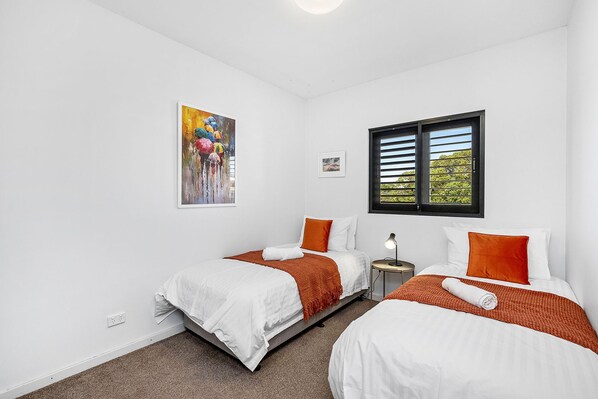 A room with two beds, white bedding, orange throw blankets, a painting, a window with blinds, and a small table with a lamp.