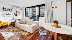A modern living room with a sofa, a dining table, and a large window with a view of a building. - Bespoke East Perth - Sleeps 2 - Superior Wifi (Perth)