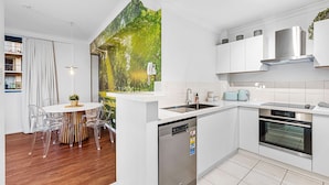 A modern kitchen with a dishwasher, oven, and white cabinetry. There is a dining area with a table and chairs. - Bespoke East Perth - Sleeps 2 - Superior Wifi (Perth)