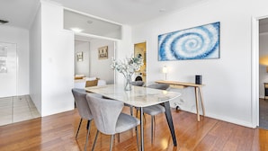 A modern dining area with a glass-top table, grey upholstered chairs, and a blue abstract painting on the wall. - Modern 2BR Pool Gym Parking (Perth)