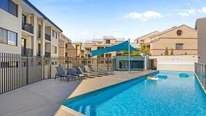 A swimming pool with lounge chairs and a shaded area, surrounded by apartment buildings. - Modern 2BR Pool Gym Parking (Perth)