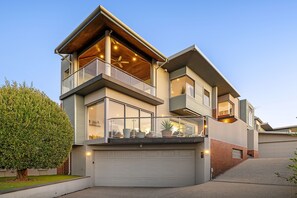 Une maison moderne à deux étages, dotée d’un garage, de grandes fenêtres et d’un balcon agrémenté de plantes en pot.