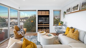 A modern living room with a sofa, armchair, and coffee table, a TV, and a large window with a view of buildings and trees. - Designer Luxury 3brd - Epic Views - South Perth (Perth)