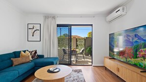 A modern living room with a blue sofa, a round coffee table, a flat-screen TV, and a sliding glass door leading to an outdoor area. - Perfect Location 2BR 1bth - Beckenham (Perth)