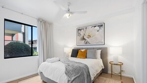 A bedroom with a large bed, a ceiling fan, a window with curtains, a framed picture on the wall, and a bedside table with a lamp. - Perfect Location 2BR 1bth - Beckenham (Perth)