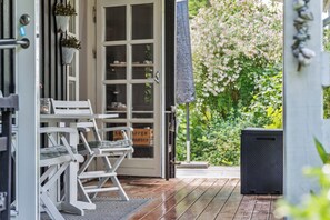 Un molo in legno con una sedia pieghevole bianca e un tavolo, una porta vetrata con cornice bianca, e un giardino fiorito.