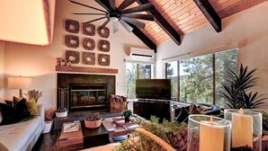 A cozy living room with a fireplace, a television, and a large window offering a view of trees.