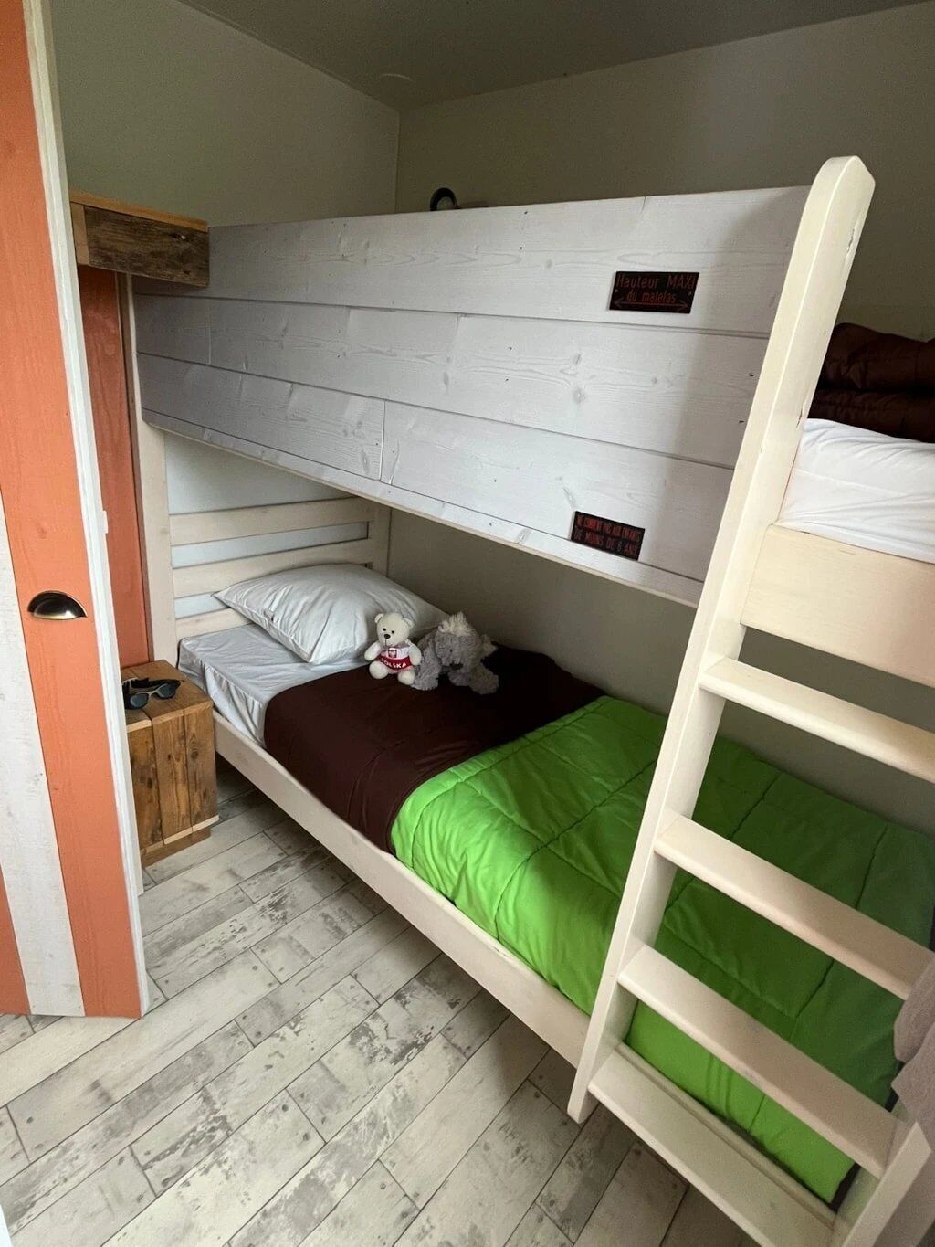 A bunk bed room with a wooden ladder, a small wooden stool, and a teddy bear on the lower bunk.