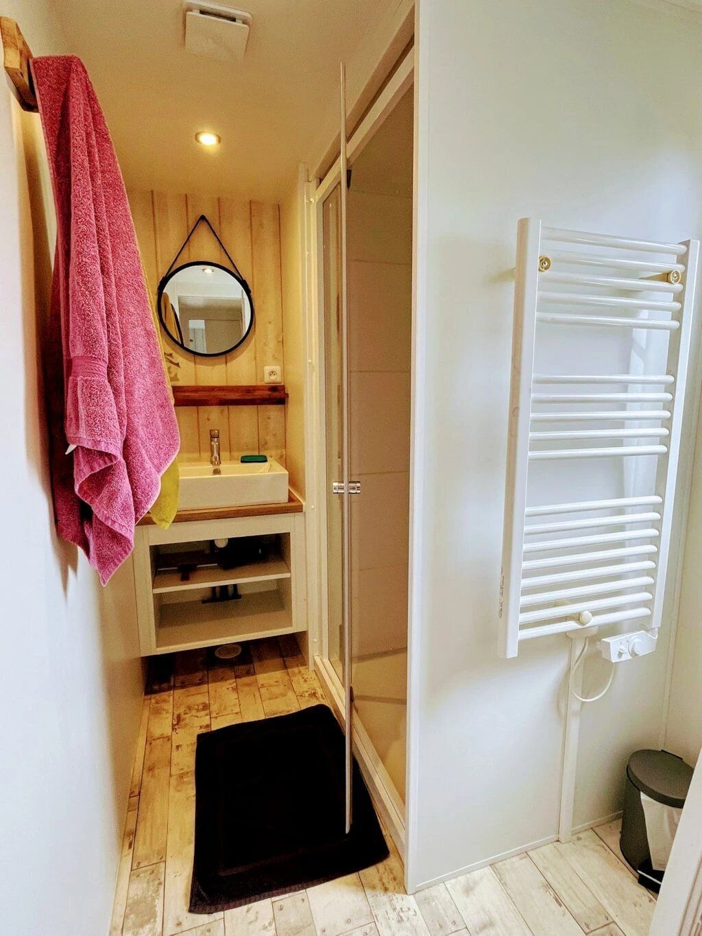 A bathroom with a wooden vanity, a mirror, a towel rack, a shower with a glass door, and a white radiator.