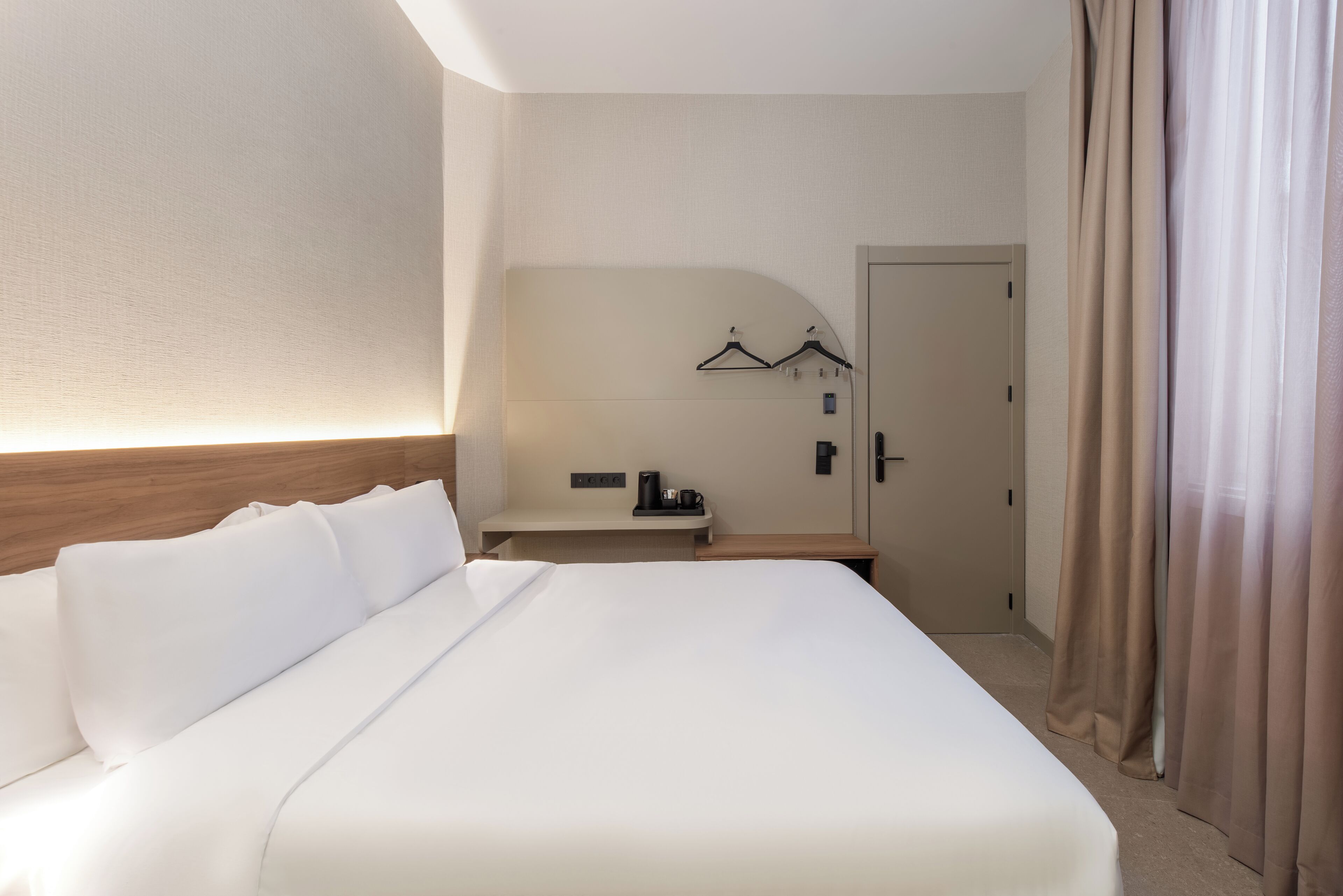 Quarto de hotel com uma cama grande, uma mesa de cabeceira com cafeteira, um porta-casacos na parede e uma porta com cortina.