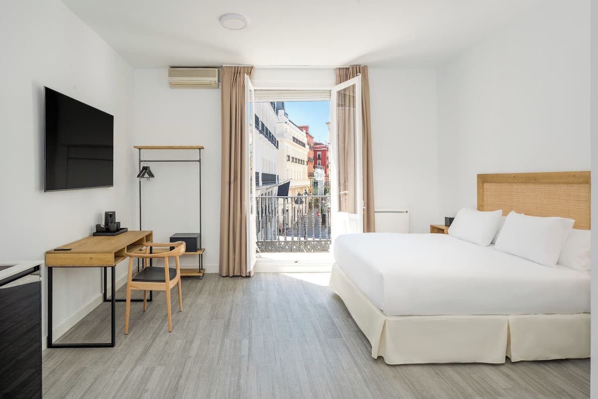 A hotel room with a bed, desk, chair, TV, and a balcony with a city view.
