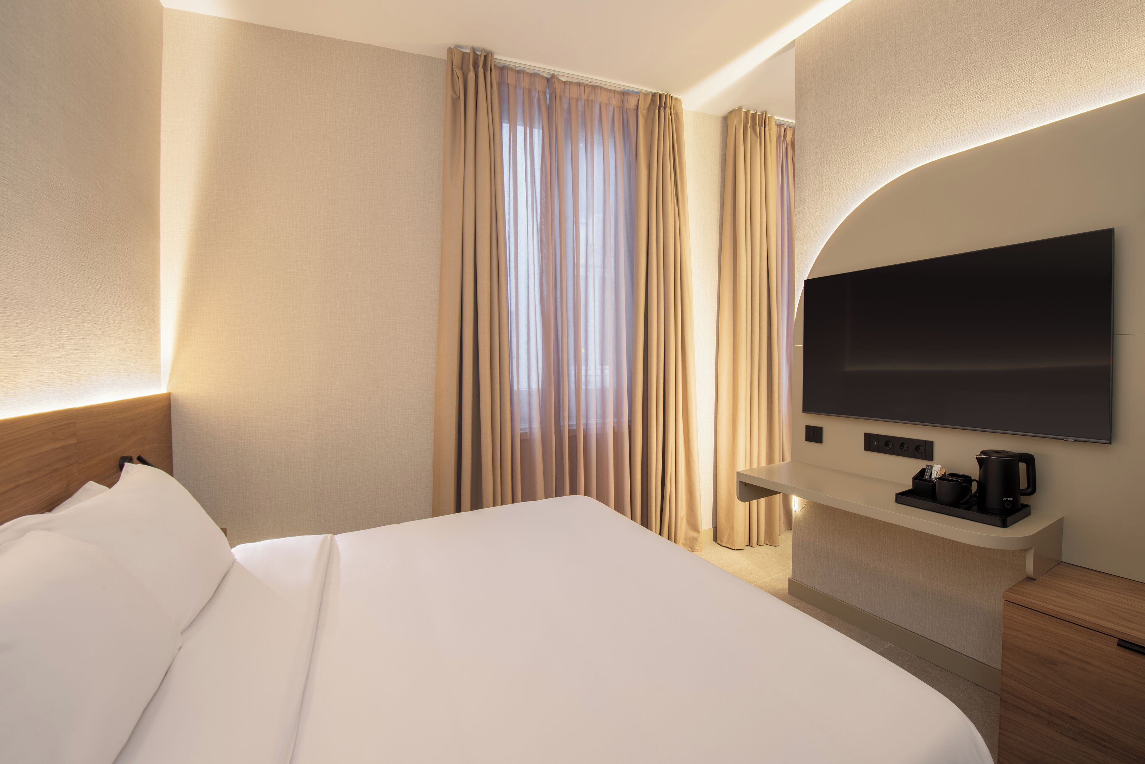 A hotel room with a large bed, a flat-screen TV, a bedside table with a coffee maker, and a window with curtains.
