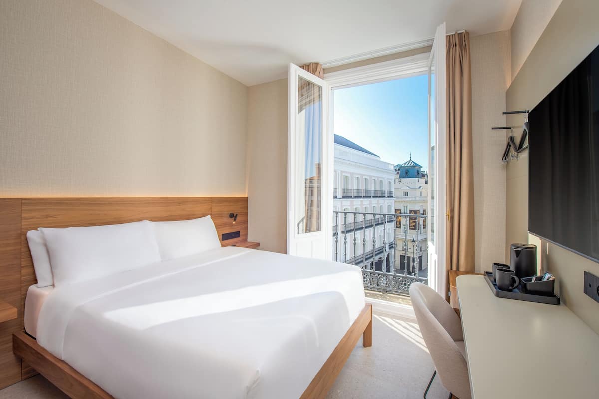 A hotel room with a large bed, a desk, and a view of the cityscape through an open door.
