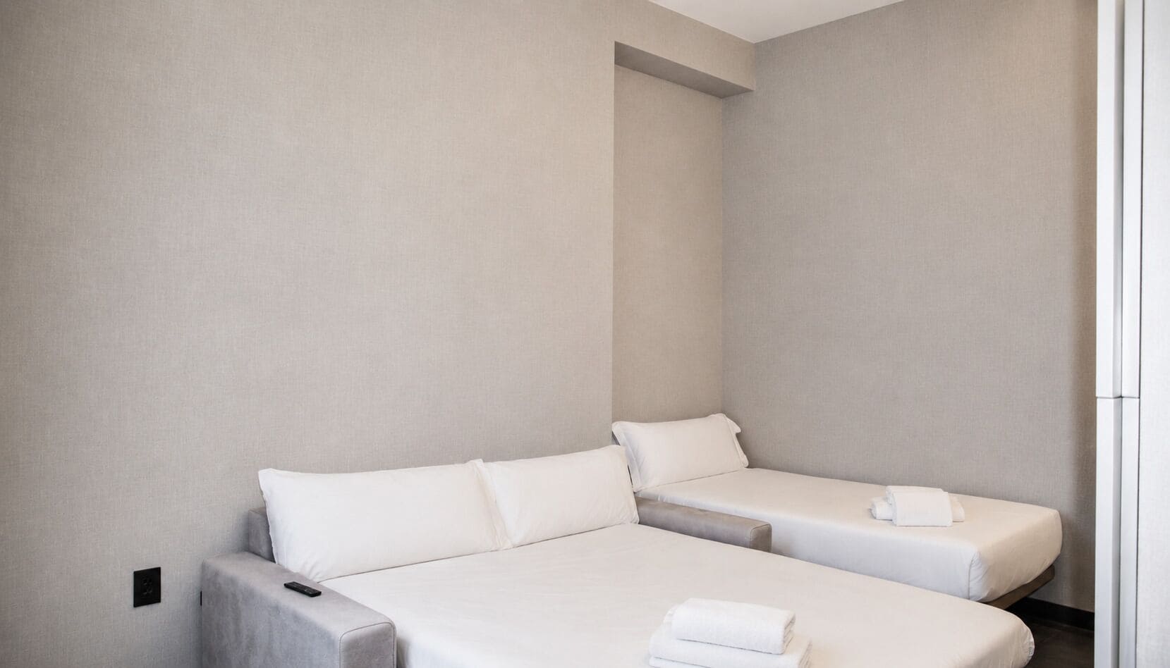 A hotel room with two beds, a small table, and a wall-mounted light.