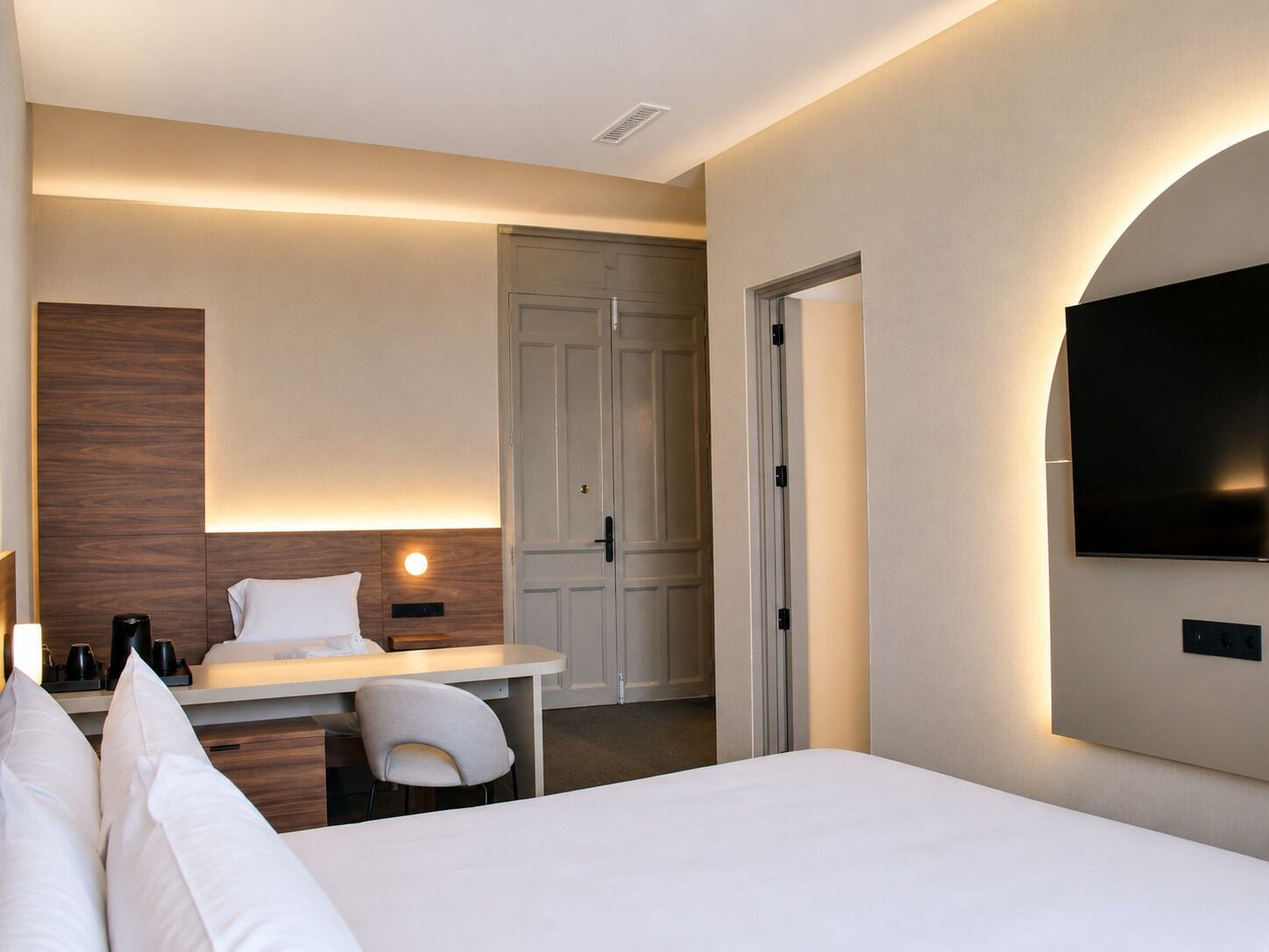 A hotel room with a bed, a desk, a chair, a TV mounted on the wall, and a door.