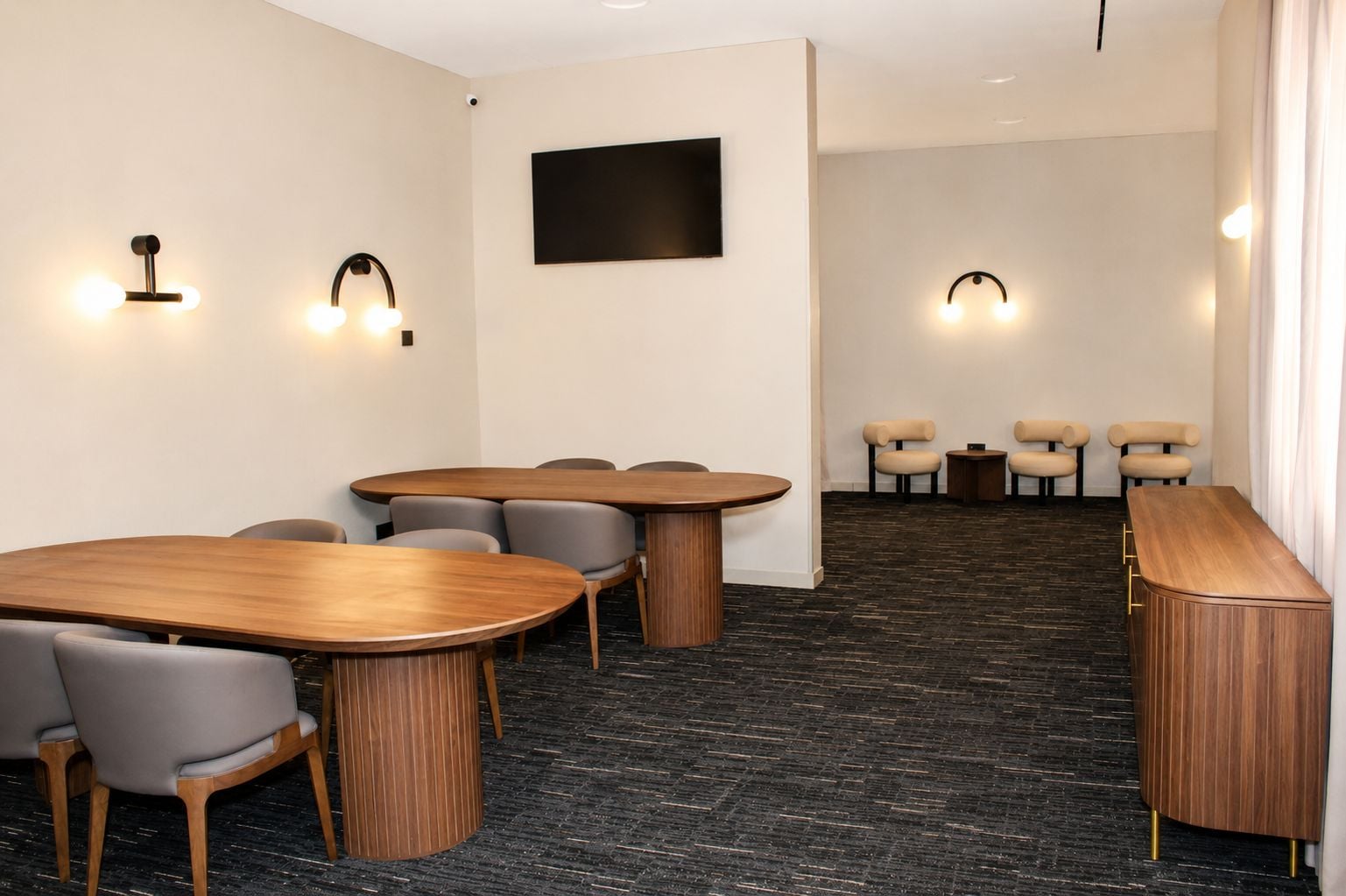 A conference room with a large oval table, several chairs, a flat-screen TV mounted on the wall, and two wall-mounted light fixtures.