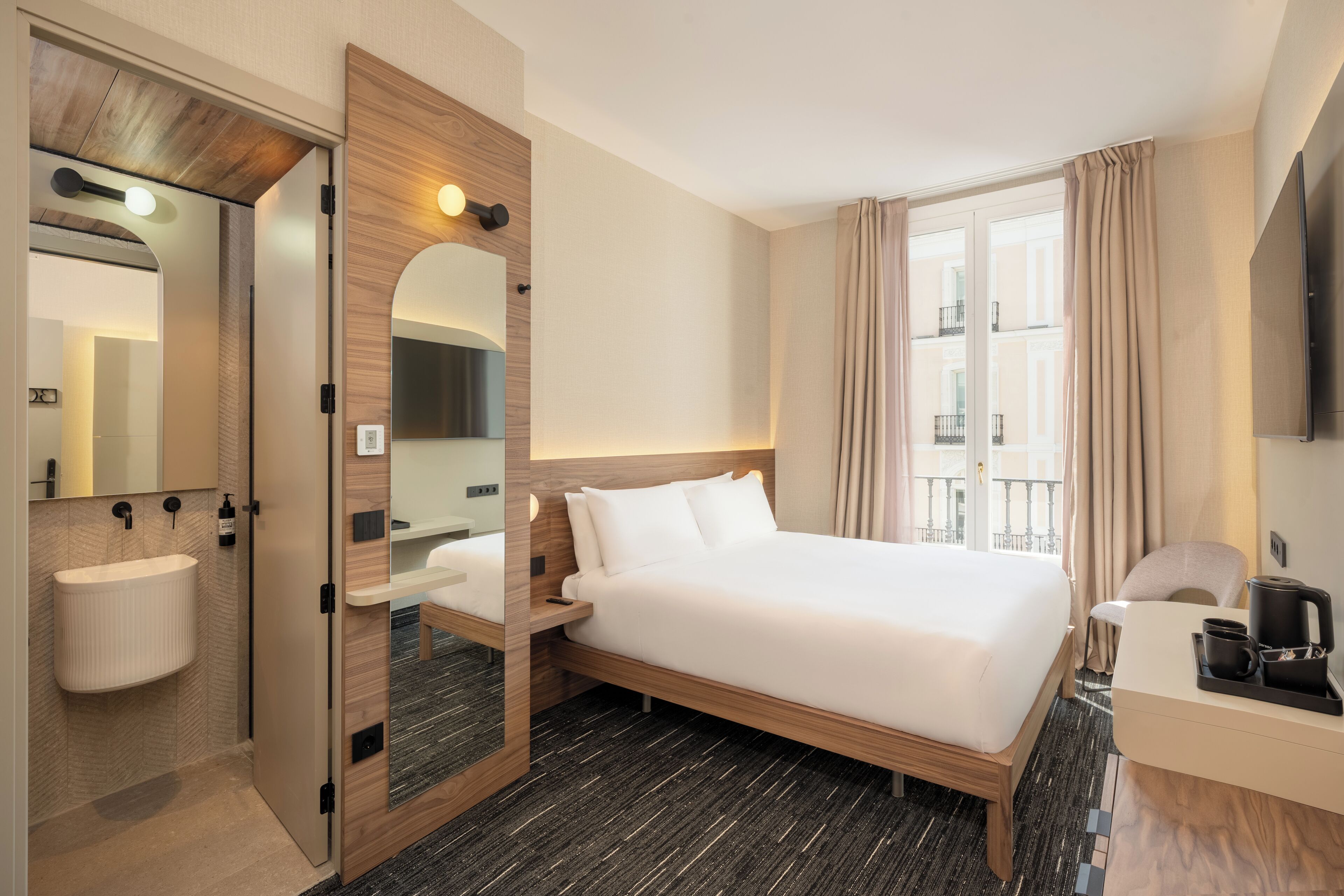 Habitación de hotel con cama, mesita de noche con cafetera, baño con lavamanos y espejo, y vista al exterior.