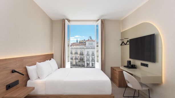 Ett modernt hotellrum med en stor säng, ett skrivbord, en platt-TV och utsikt över staden genom ett stort fönster.