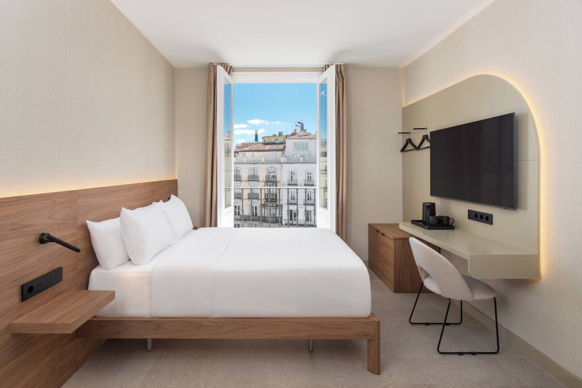 A modern hotel room with a large bed, a desk, a flat-screen TV, and a view of the city through a large window.