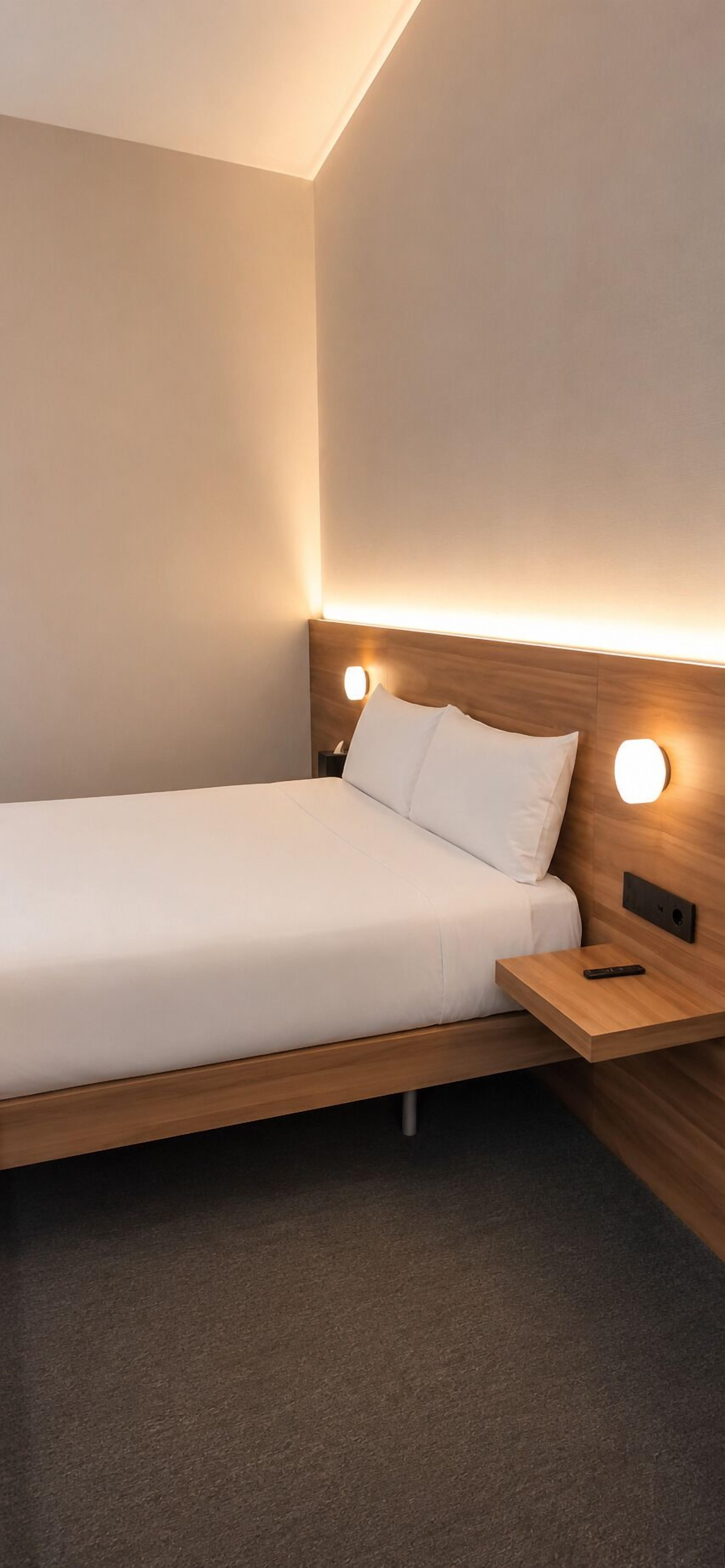 A single bed with a wooden headboard, a bedside table with a remote control, and a wall-mounted light fixture.