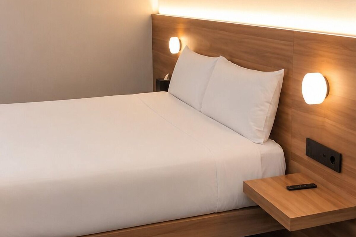 Een enkel bed met een houten hoofdbord, een nachtkastje met een afstandsbediening en een wandlamp.