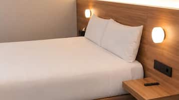 A single bed with a wooden headboard, a bedside table with a remote control, and a wall-mounted light fixture.