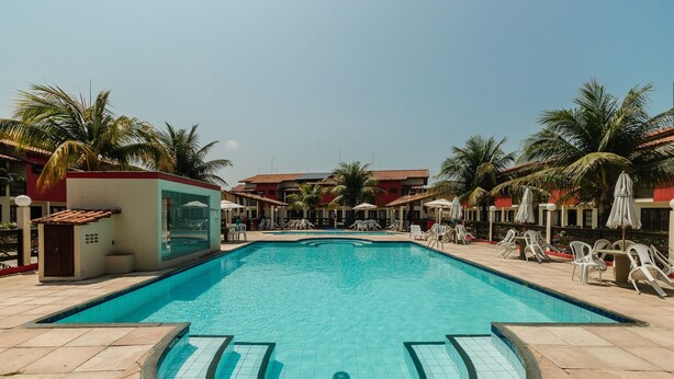 Ein Swimmingpool mit Fliesenrändern, umgeben von Liegest ühlen und Sonnenschirmen, mit Palmen und Gebäuden im Hintergrund.