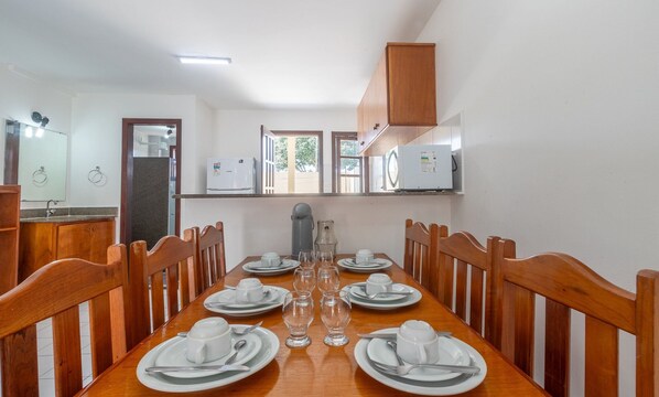 A dining area with a wooden table set for six, surrounded by wooden chairs. A refrigerator and a coffee maker are visible in the background.