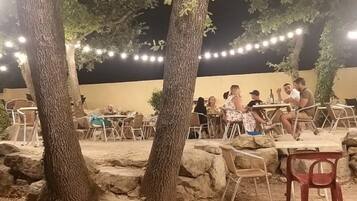 Un espace extérieur aménagé avec des tables et des chaises, entouré de rochers et d’arbres, et illuminé par des guirlandes lumineuses.