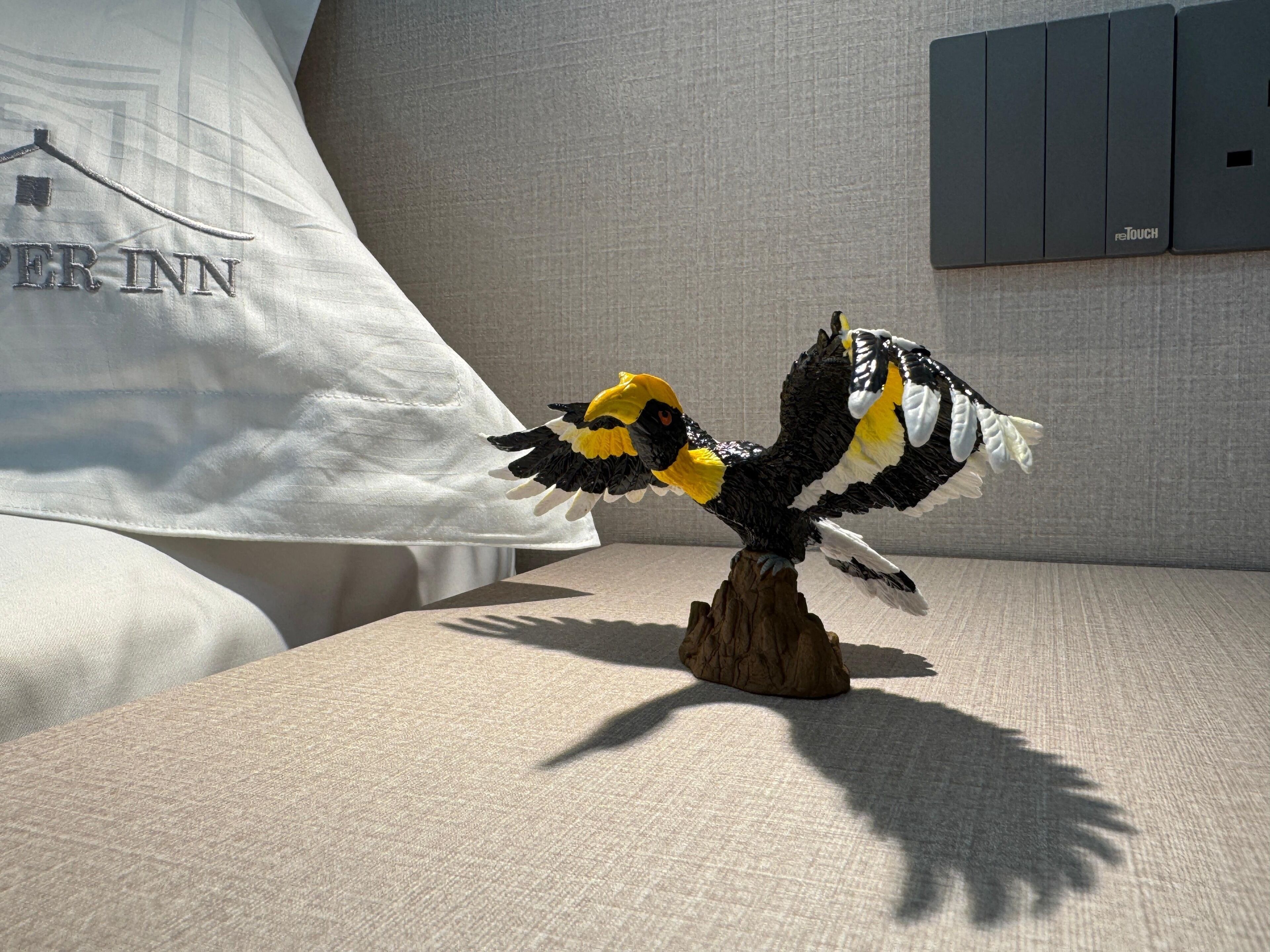 Eine Vogel-Figur auf einer strukturierten Oberfläche, mit einem Kissen und einer Wand im Hintergrund.