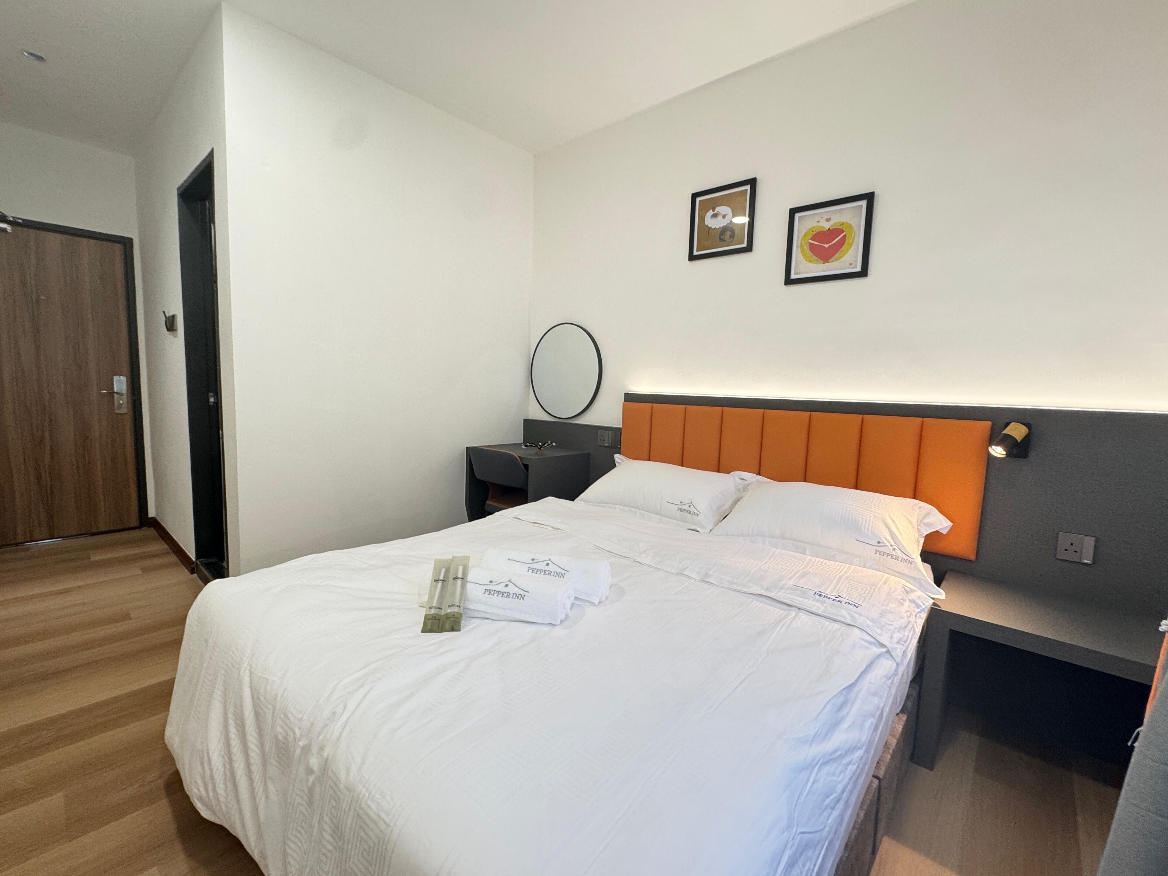 Una cama bien hecha con sábanas blancas y un almohadón con un logotipo, una mesita de noche con una lámpara, y dos cuadros enmarcados en la pared.