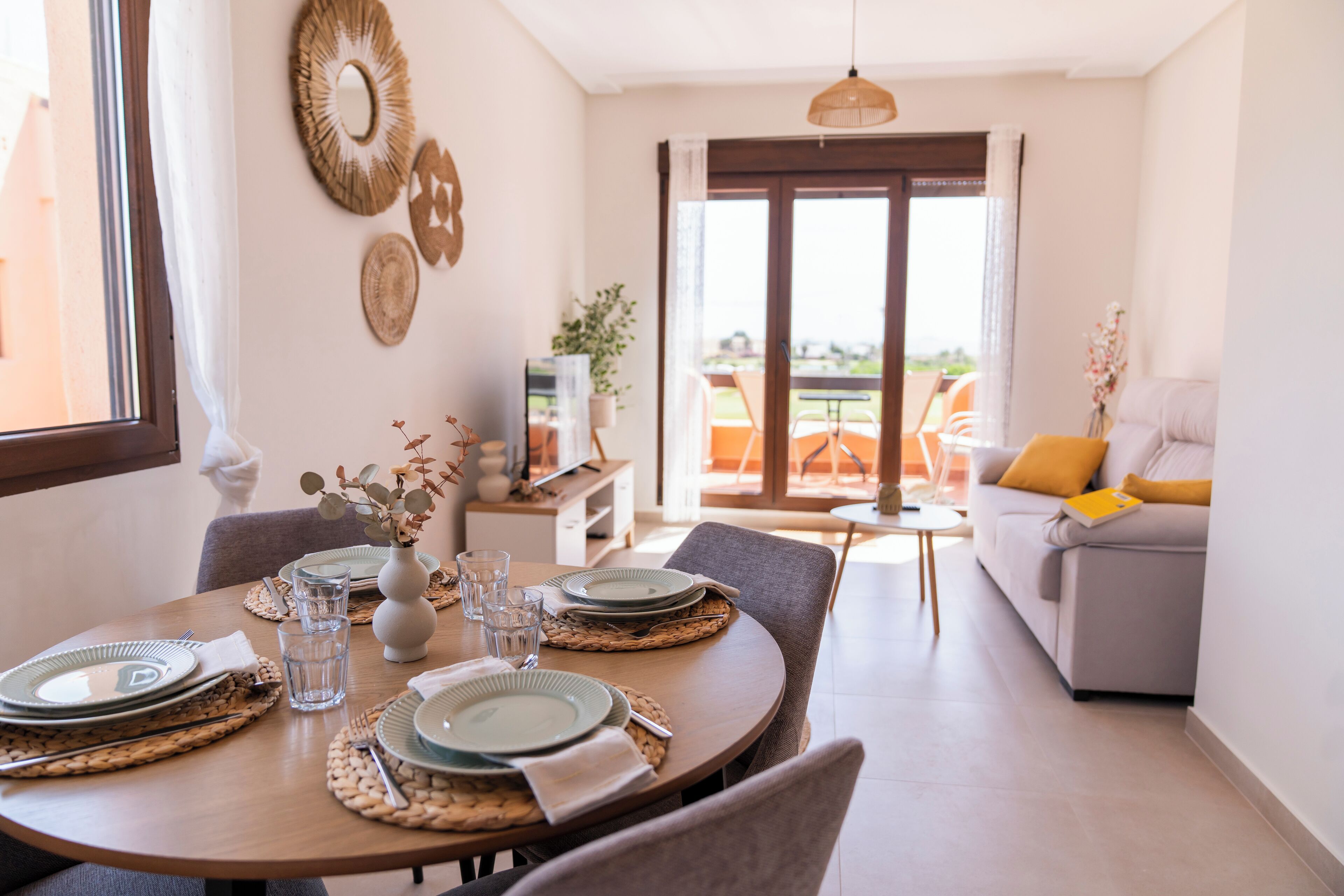 A dining area with a round table set for four, a sofa with a yellow pillow, and a balcony with orange chairs.