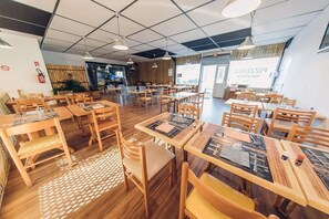 A restaurant with wooden tables and chairs, a tiled ceiling, and a visible kitchen area.