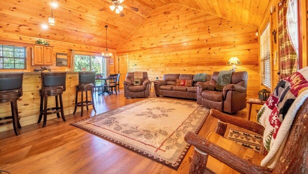 A cozy cabin interior with wooden walls and floors, a central seating area with a sofa and chairs, a bar area with stools, and a large rug. - *Amazing 2BD Cabin Near TOWN! Hot Tub + Fire Pit* (Sevierville)