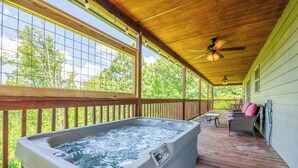 A hot tub on a wooden deck with a view of trees, a ceiling fan, and a patio table. - *Amazing 2BD Cabin Near TOWN! Hot Tub + Fire Pit* (Sevierville)