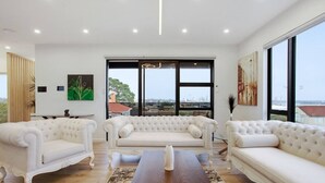 A spacious living room with a large window, white sofas, a wooden coffee table, and a view of the city.