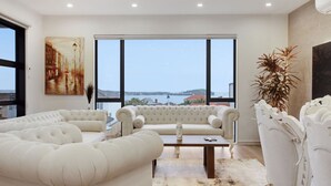 A spacious living room with a large window offering a view of the sea and buildings. There are white upholstered sofas and a coffee table.