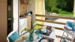 A dining area with a table set for two, a kitchen area with cabinets and a refrigerator, and a balcony with a view of greenery.