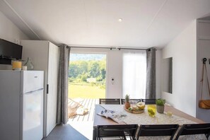 Cozinha moderna com geladeira, mesa de jantar com cadeiras, televisão e vista para uma paisagem verde através de uma porta.