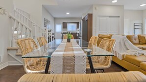 Ein modernes Wohnzimmer mit einem Esstisch mit Glasplatte, Korbstühlen, einem gelben Sofa und einer Treppe mit weißen Geländern.