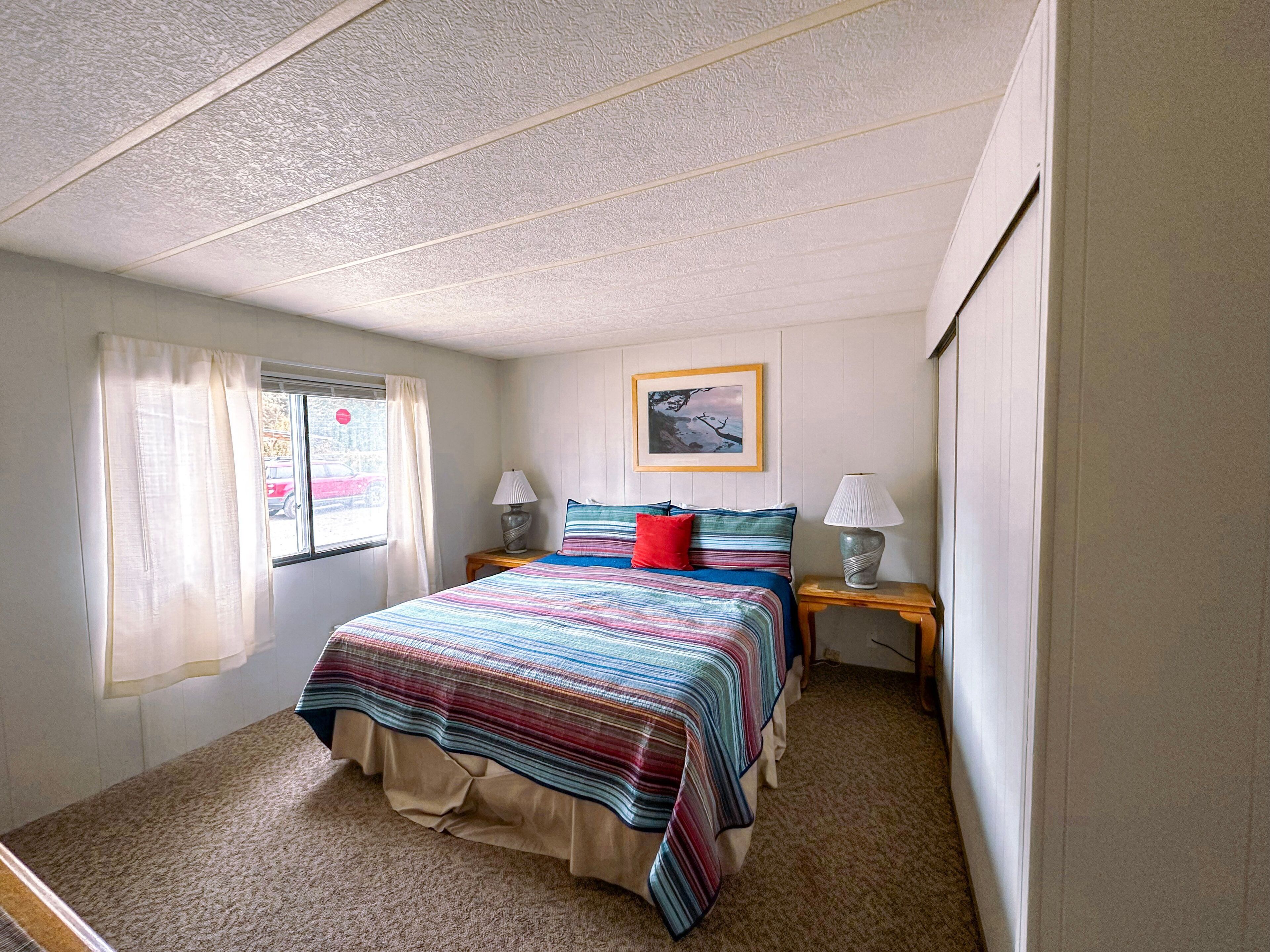 Un dormitorio con una cama, dos mesitas de noche, una ventana con cortinas e imagen enmarcada en la pared.