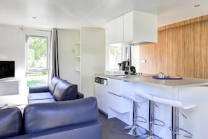 A compact kitchen with white cabinets, a built-in oven, and a coffee machine. There is a bar area with stools and a television mounted on the wall.