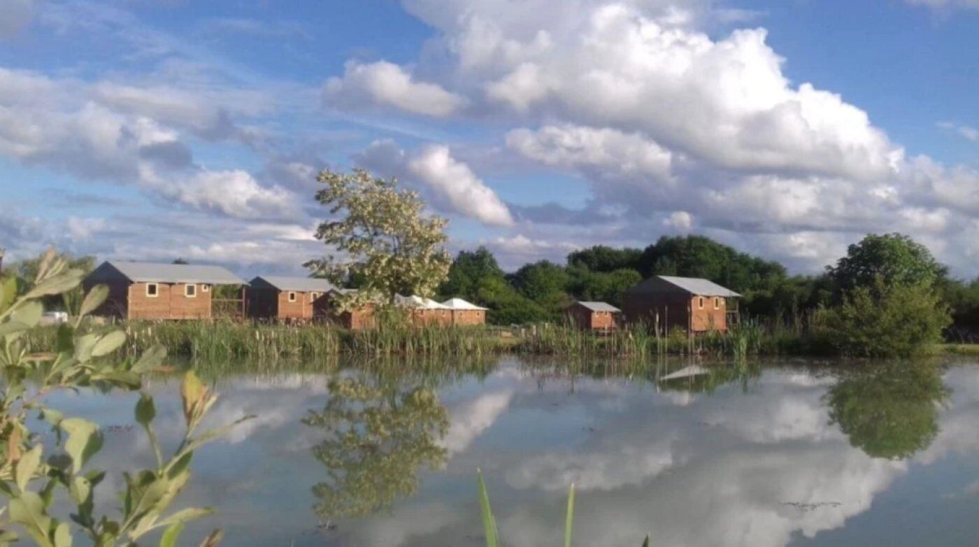 Filas de cabañas de madera junto a un lago tranquilo, con reflejos del cielo y los árboles.