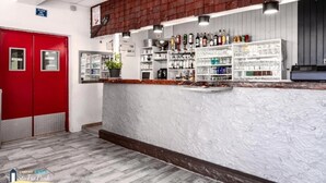 Zona de bar con estantes llenos de botellas y vasos, una puerta roja y una pared blanca con textura.