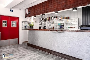 Una zona de bar con estanterías llenas de botellas y vasos, una puerta roja y una pared blanca con textura.