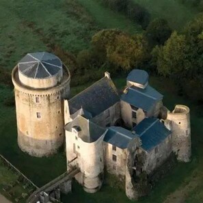 A historic stone building with a round tower and multiple connected structures, surrounded by greenery.