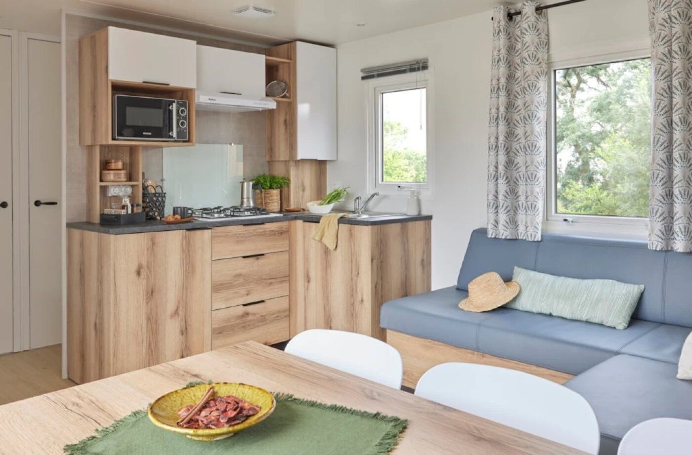 Une cuisine compacte avec des meubles en bois, un four à micro-ondes et un évier. On y trouve une table à manger avec un bol de nourriture et un set de table vert. Un canapé bleu, agrémenté d’un chapeau et d’un coussin, est placé devant une fenêtre ornée de rideaux à motifs.