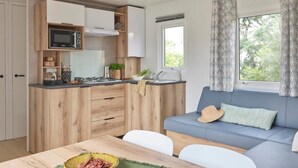 Une cuisine compacte avec des meubles en bois, un four à micro-ondes et un évier. On y trouve une table à manger avec un bol de nourriture et un set de table vert. Un canapé bleu, agrémenté d’un chapeau et d’un coussin, est placé devant une fenêtre ornée de rideaux à motifs.