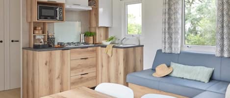 Een kleine keuken met houten kastjes, een magnetron en een spoelbak. Er staat een eettafel met een schaal eten en een groene placemat. Een blauwe bank met een hoed en een kussen staat voor een raam met decoratieve gordijnen.